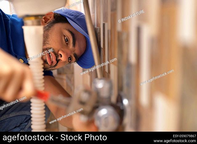 A YOUNG PLUMBER LEANING DOWN AND FIXING LEAKAGE OF A PIPE