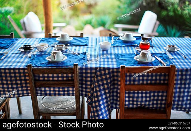 Table with empty plates and coffee cups, blue chequered tablecloth, ready for morning breakfast, blurred green foliage in background - eating at tropical...
