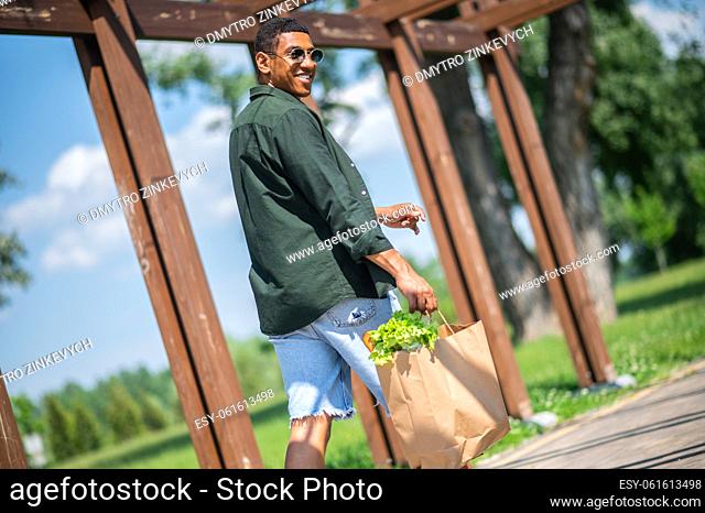 Optimism. Dark-skinned attractive guy in sunglasses smiling looking back at camera walking in park holding food bag at warm day