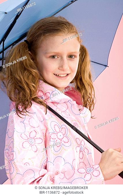 Red-haired girl holding a light blue umbrella and wearing a raincoat in front of a pink backdrop