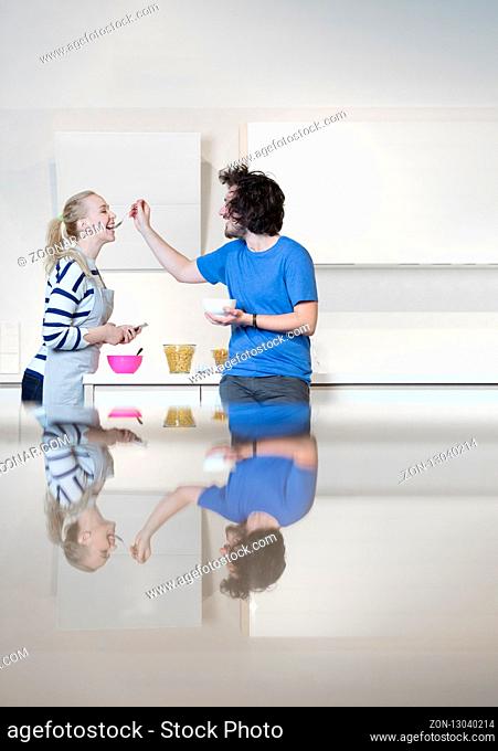 Beautiful young couple is feeding each other and smiling while cooking in kitchen at home