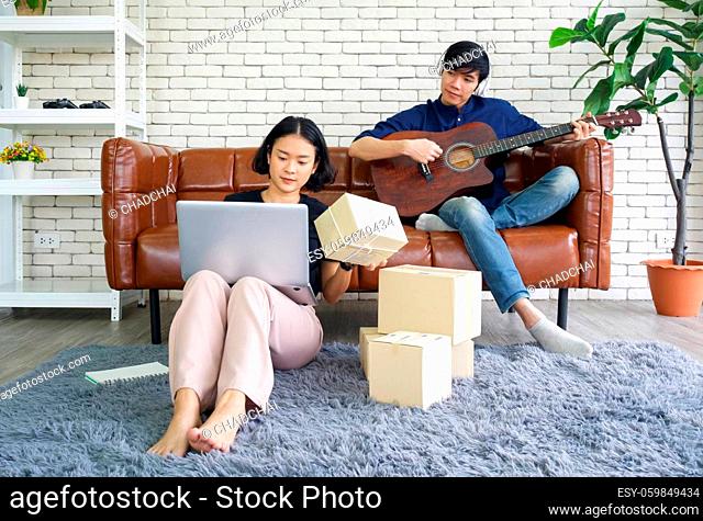 Young lovers spend time together on holidays in the living room. Asian man with headphones playing an acoustic guitar while the girl check the parcel delivery...
