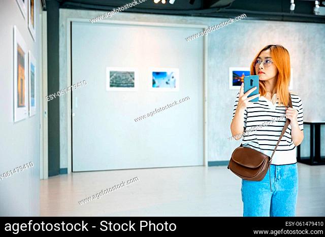 Visitor woman standing takes picture art gallery collection in front framed paintings pictures on white wall with mobile phone