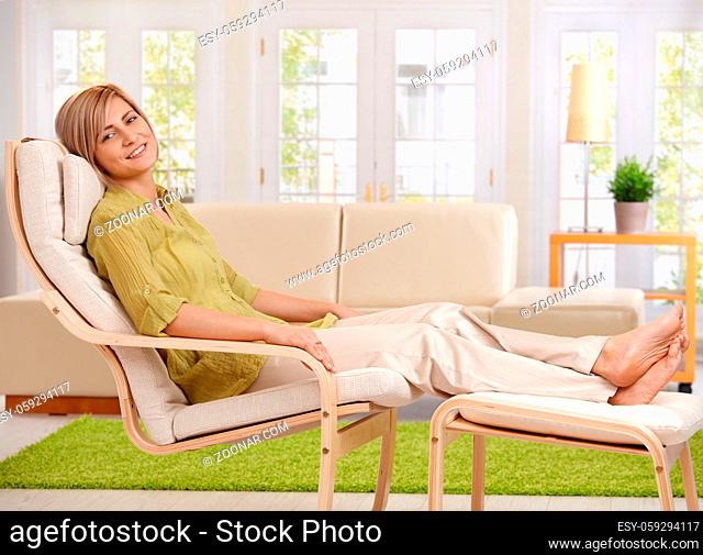 Woman relaxing at home, sitting in armchair with crossed feet up on footboard, smiling at camera