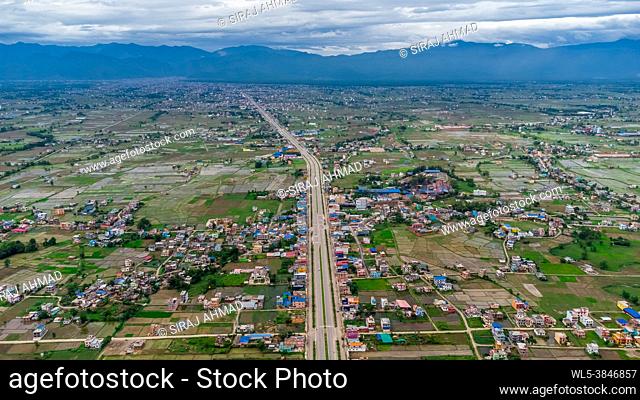 Aerial view of six-lane highway passing through meadows, fields and scattered houses towards mountains in countryside Nepal