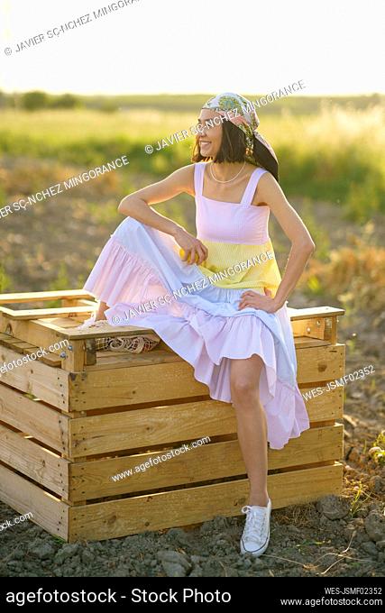 Smiling young woman sitting on wooden crate while looking away in the countryside