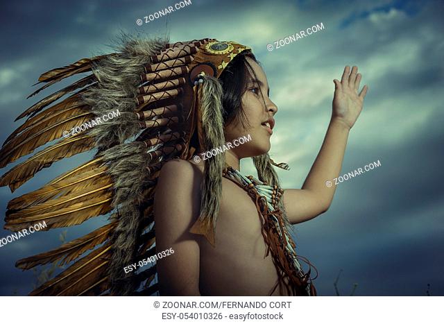 Child dressed as an American Indian at sunset, wears a plume of Indian feathers and breastplate of bones. he has raised his hand as a greeting to nature