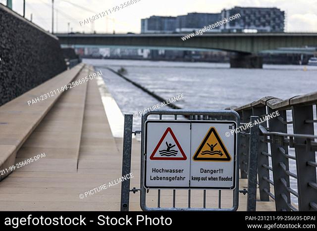 11 December 2023, North Rhine-Westphalia, Cologne: Signs with the words ""Flood - danger to life"" hang on the partially flooded flight of steps on the banks of...
