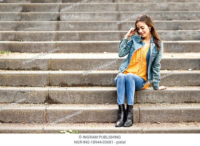 Young woman with nice hair wearing casual clothes in urban background. Happy girl with wavy hairstyle sitting in urban stairs