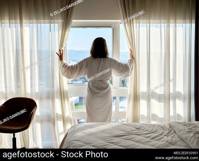 Woman opening white curtain while looking through window at luxury hotel room