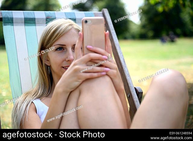 Young blonde woman in London Hyde Park, sitting in a beach chair and enjoying the late summer sun and browsing the internet on her mobile phone
