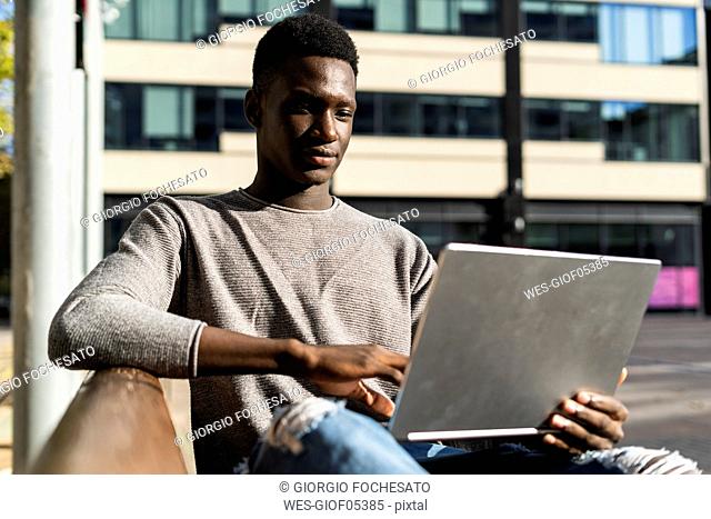 Young man sitting on a bench in the city, using laptop