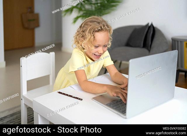 Happy blond girl using laptop sitting at table in living room