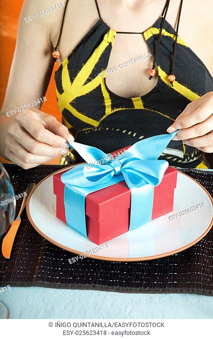 woman with brown and yellow dress opening blue bow of gift red present box with hands, over white dish in table of restaurant