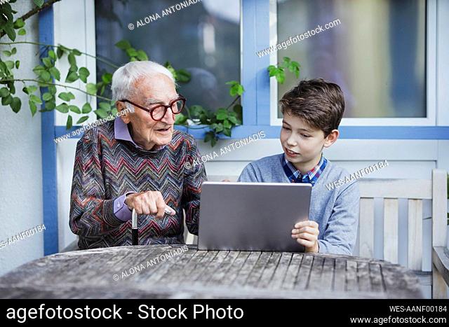 Senior man talking to grandson sitting with tablet PC at table