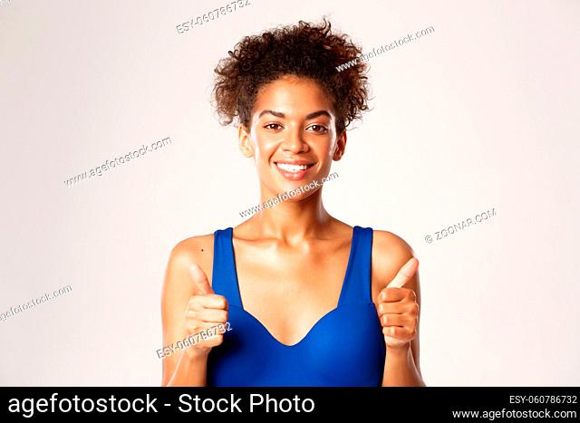 Attractive and healthy african-american fitness woman in blue sports bra, showing thumbs-up in approval and smiling, standing over white background