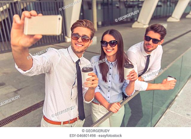 Handsome young businessmen and lady in sun glasses are holding coffee, making selfie using smart phone and smiling, standing on balcony of the office building