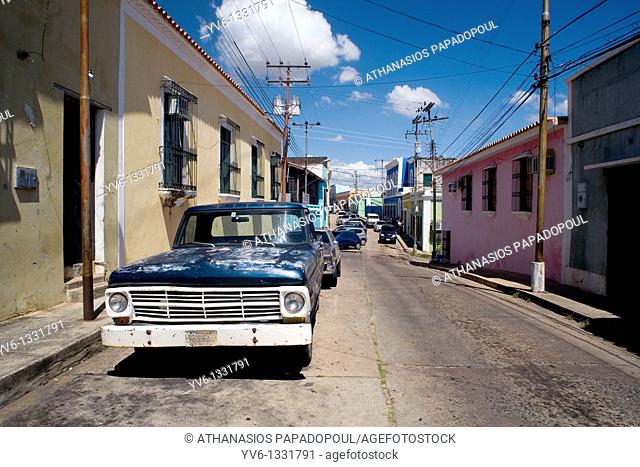 Photograph Of A Typical Street At Ciudad Bolivar Historical Center With An Old American Car At The Front Of The Image And Colored Houses And Other cars Shoot On...