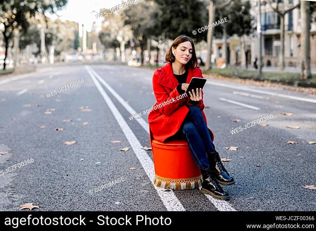 Young woman in red jacket listening music while using digital tablet sitting on seat at street