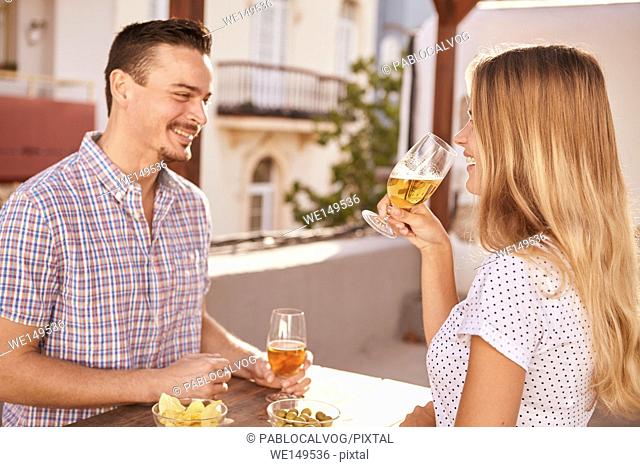 Happily smiling couple having some drinks while sitting across from each other at a table with bowls of snacks in bright sunlight