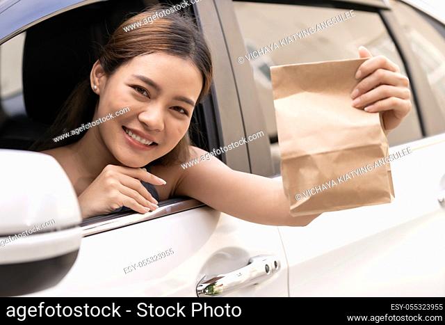 Asian Young adult in car holding disposable bag for take out food from drive thru service restaurant. Drive thru is new normal and popular service after...