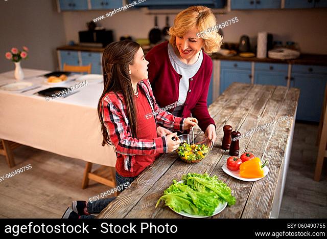 Grandma and granddaughter standing at big wooden table. Girl mixing salad