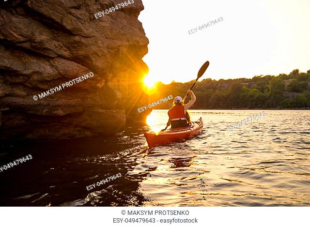 Young Woman Paddling the Red Kayak on the Beautiful River or Lake near High Rocks at Sunset