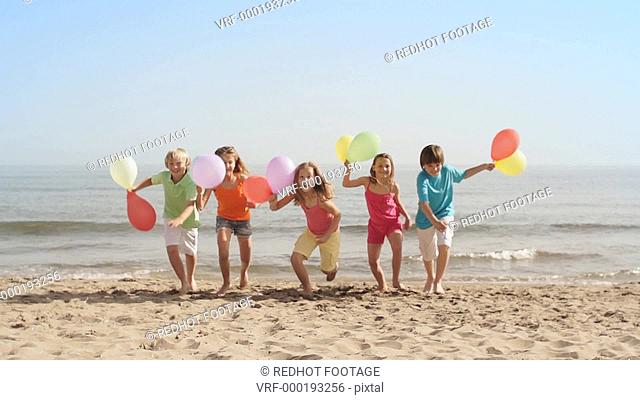 Slow motion of five children running towards camera on beach holding balloons