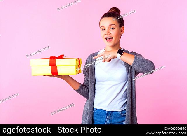 Birthday surprise, present. Portrait of happy teenage brunette girl with bun hairstyle in casual clothes pointing at gift box and smiling joyfully at camera