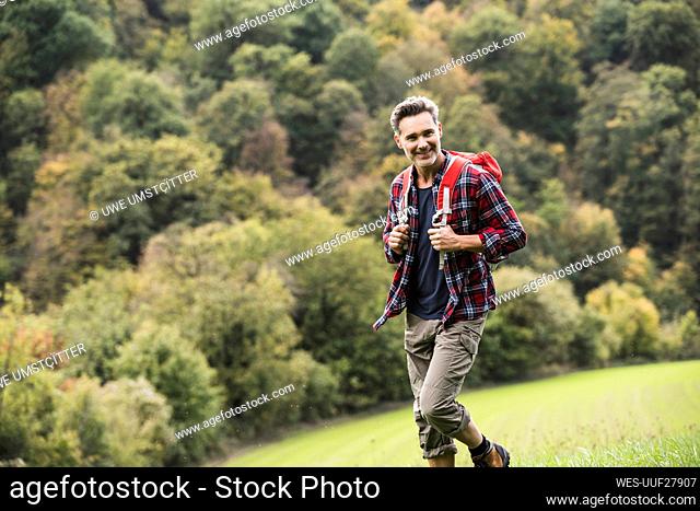 Smiling man with backpack hiking on grass in front of trees