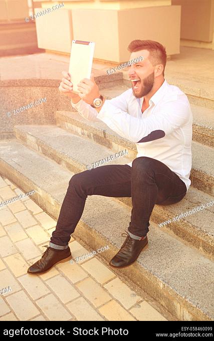 Young man using tablet computer for making selfies. Bearded man in white shirt and black jeans smiling for the camera outdoors. Toned image