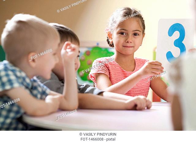 Portrait of cute girl holding number 3 placard while sitting beside friends in classroom
