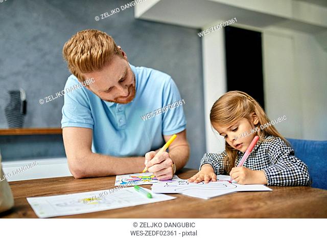 Father and daughter sitting at table, painting colouring book