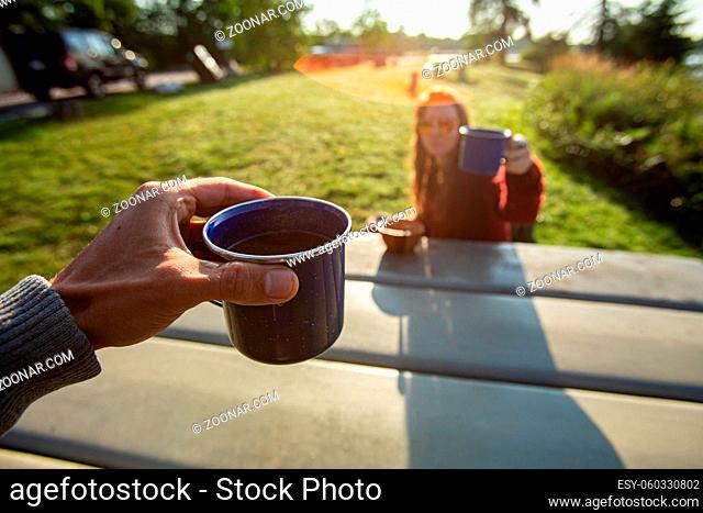 Selective focus point of view photo of two people cheering their cups of coffee while enjoying a nice outdoor picnic in a nature environment