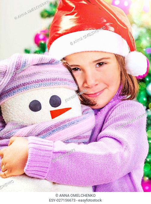 Closeup portrait of a cute little baby girl wearing red Santa hat near festive decorated Christmas tree, with love hugging her soft snowman toy
