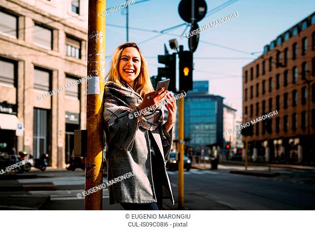 Young woman with smartphone laughing at tram station, Milan, Italy