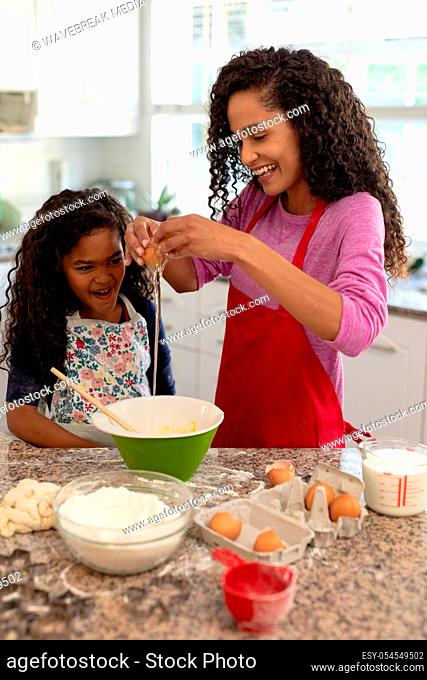 Front view of a mixed race woman in a kitchen with her young daughter at Christmas, making cookies cracking an egg