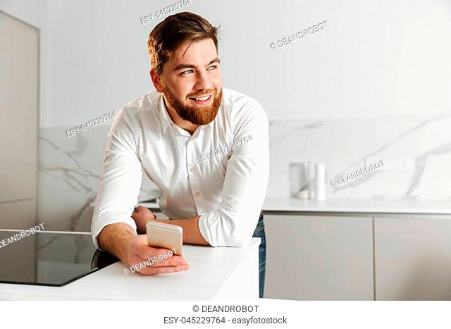 Portrait of a happy young businessman dressed in white shirt holding mobile phone while leaning on a table indoors