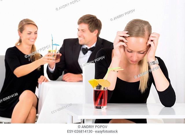 Depressed Woman Sitting In Restaurant In Front Of Couple Toasting Glass Of Cocktails