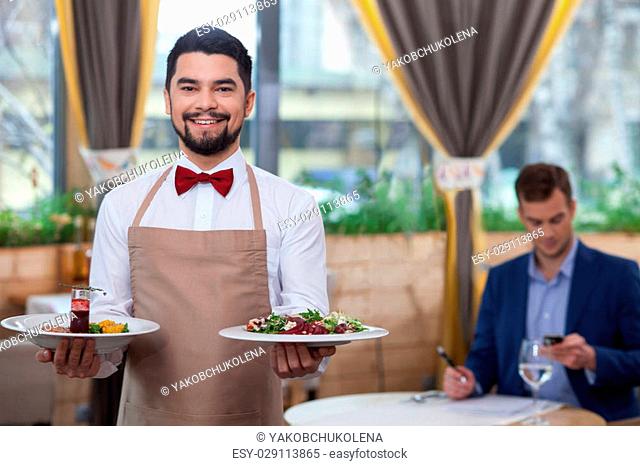 Handsome male cafe worker is serving a customer. He is holding plates of salad and smiling. The man is standing and looking at camera with joy
