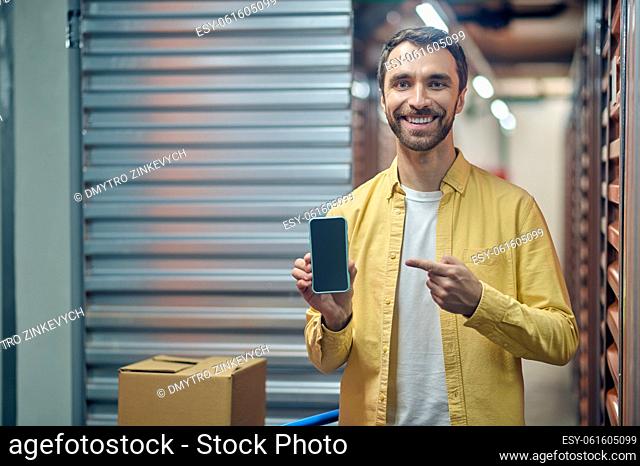 Waist-up portrait of a smiling cheerful storehouse employee pointing at the smartphone in his hand