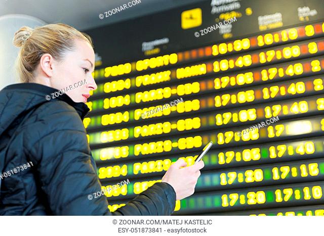 Young woman at international airport looking at the flight information board, holding smart phone in her hand, checking flight informations on phone application
