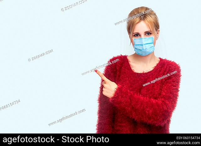 Portrait of happy beautiful young woman with surgical medical mask in red blouse standing, looking at camera and pointing at background copyspace