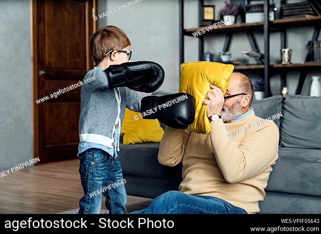 Playful boy wearing boxing gloves playing with grandfather covering face with pillow in living room