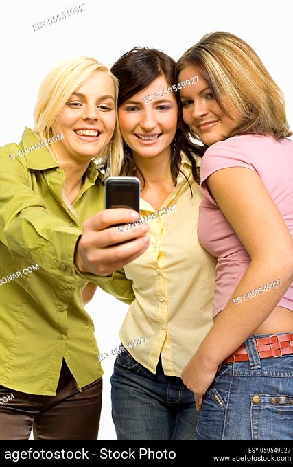 Three girl friends are standing. One of them is holding mobile phone. She's showing its screen to her friends. Isolated on white in studio
