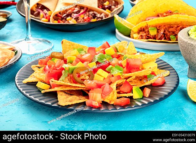 Nachos with avocado and tomato, with jalapeno peppers and cilantro leaves, on a table with other Mexican food