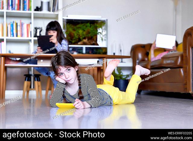 Little girl with smartphone lying on the floor at home while her sisters in the background using digital tablets