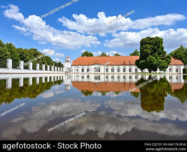 05 July 2022, Brandenburg, Neuhardenberg: An outbuilding of Neuhardenberg Castle and the Schinkel Church in the background are reflected in the water of a pond
