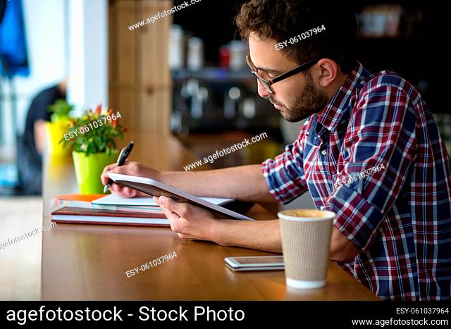 Handsome student man looking at tablet PC and writing essay or publication with hand. Man in glasses studying in restaurant