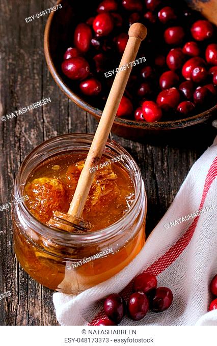 Open glass jar of liquid honey with honeycomb and honey dipper inside and fresh cranberries in vintage bowl over old wooden table with white kitchen towel